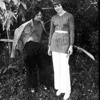 Two women stand outdoors in a leafy, wooded area.