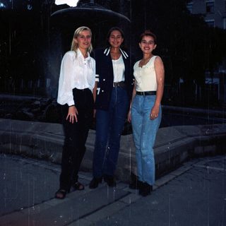 Three women stand outdoors in casual, 90s-era clothing.
