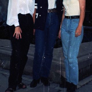 Three women stand outdoors in casual, 90s-era clothing.