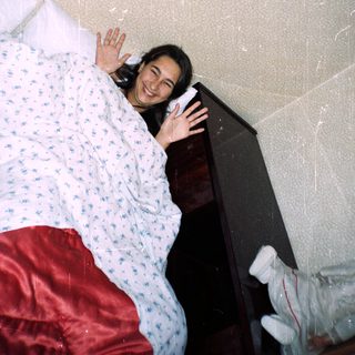 Person lying on a bed waving, teddy bear visible.