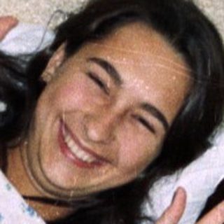 Person lying on a bed waving, teddy bear visible.