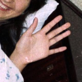 Person lying on a bed waving, teddy bear visible.