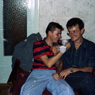 Two men sit on a couch, sharing a cup.