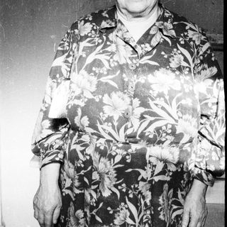 Here's a brief summary of the image: Elderly woman stands smiling in a floral print dress.