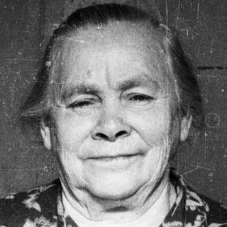 Here's a brief summary of the image: Elderly woman stands smiling in a floral print dress.