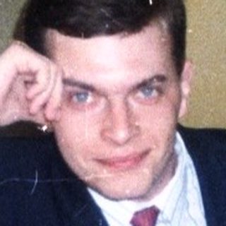 Man in suit sits on a patterned couch indoors.