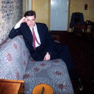 Man in suit sits on a patterned couch indoors.