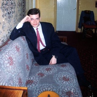 Man in suit sits on a patterned couch indoors.