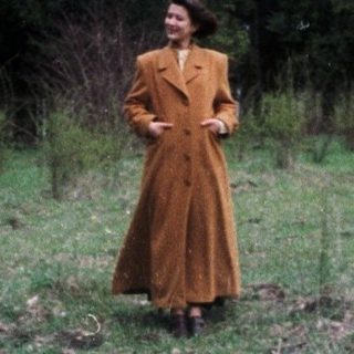 Woman in coat stands in a field, trees in background.