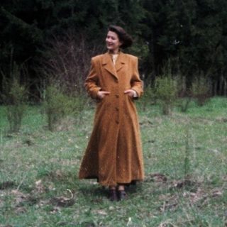 Woman in coat stands in a field, trees in background.