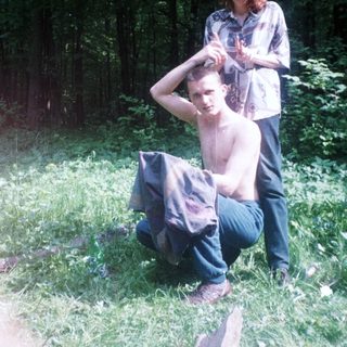 Man squats in grass; woman stands nearby in woods.