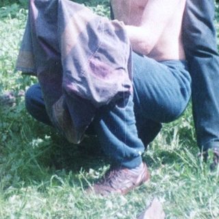Man squats in grass; woman stands nearby in woods.