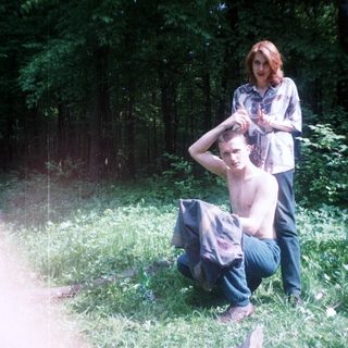 Man squats in grass; woman stands nearby in woods.