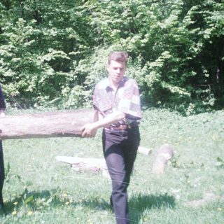 Two men carry a log in a grassy, wooded area.