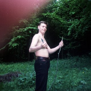 Man stands shirtless in woods, holding something, sun flares.