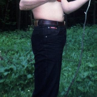 Man stands shirtless in woods, holding something, sun flares.