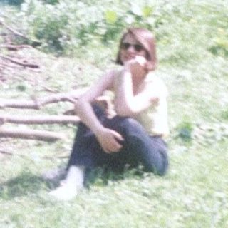 Person sits near a fire in a forest clearing.
