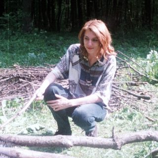Woman sits near a campfire in a wooded area.