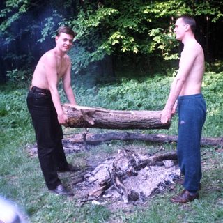 Two shirtless men carry a log near a campfire.