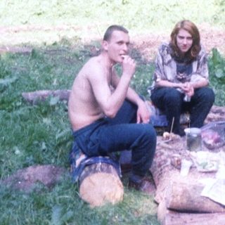 Three people relaxing outdoors on logs in a wooded area.