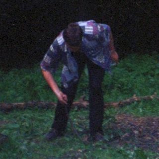 Two people near a fire in a dark, wooded area.