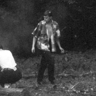 Two people near a fire in a dark, wooded area.