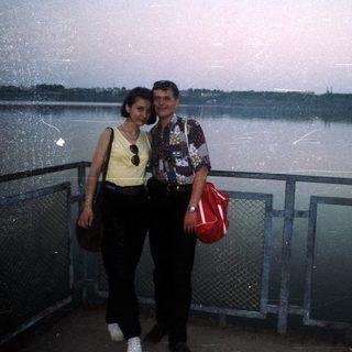 Couple poses by water with cityscape in the background.