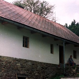 Man sits outside stone and stucco house with red roof.