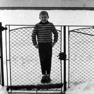 Boy stands in snow, framed by a gate.
