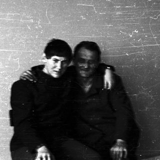 Two men sit close, appearing somber, in a room.