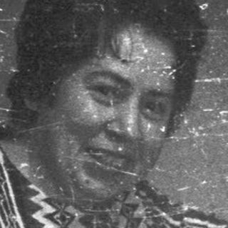 Woman in patterned dress gesturing, men seated in a room.