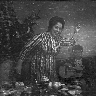 Woman in patterned dress gesturing, men seated in a room.
