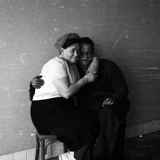 Here's a brief summary of the image: Two people embrace, seated against a stark wall.