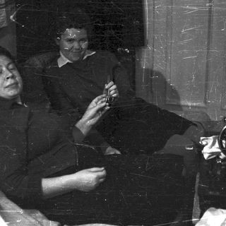 Man in suit holding object, woman seated in room.