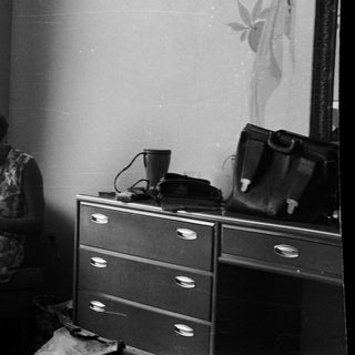 Woman seated, knitting in a room with a dresser.