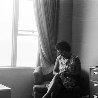 Woman seated, knitting in a room with a dresser.