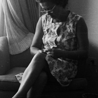 Woman seated, knitting in a room with a dresser.