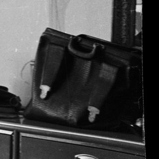 Woman seated, knitting in a room with a dresser.