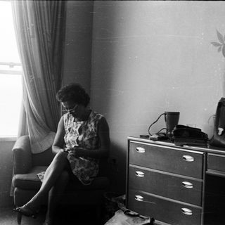 Woman seated, knitting in a room with a dresser.