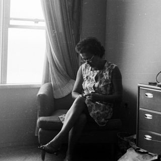 Woman seated, knitting in a room with a dresser.