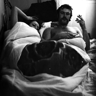Couple relaxing in bed, man smoking, guitar nearby.