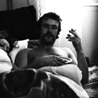 Couple relaxing in bed, man smoking, guitar nearby.