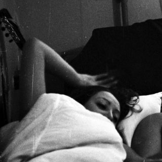 Couple relaxing in bed, man smoking, guitar nearby.