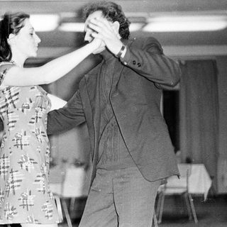 Couple dancing in a room with tables and chairs.