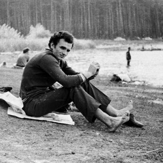Man seated on a blanket by a lake shoreline.