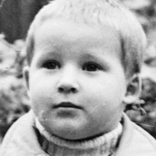 Boy in coat stands in a yard, looking forward.