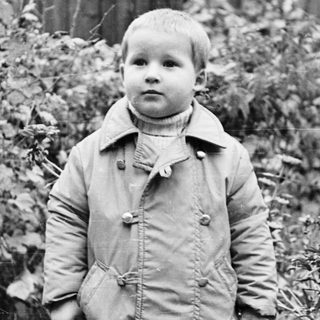 Boy in coat stands in a yard, looking forward.
