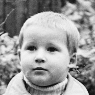 Boy in coat stands in a yard, looking forward.