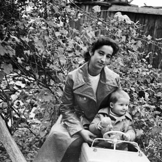 Woman and child sit on toy car near a fence.