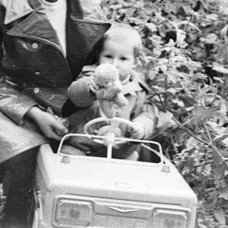 Man and child seated in a toy car outdoors.